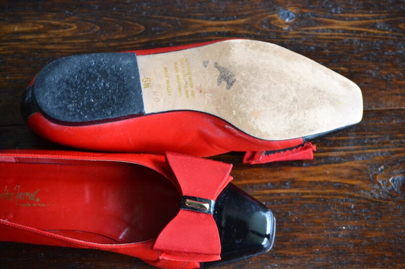 May include: Red leather ballet flats with black patent leather toe caps and a red bow detail. The sole is visible, showing wear and the size 5M. The shoes are on a wooden surface.