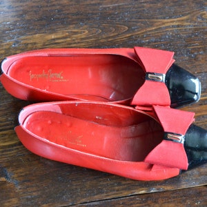 May include: A pair of red leather ballet flats with black patent leather toes and red bows. The shoes have the brand name "Jacqueline Ferrar" printed in gold inside. The shoes are on a wooden surface.