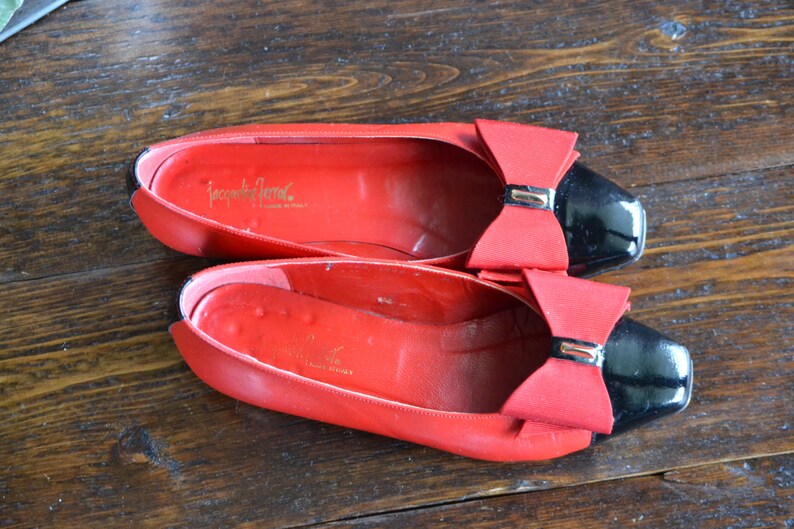 May include: A pair of red leather ballet flats with black patent leather toes and red bows. The shoes have the brand name "Jacqueline Ferrar" printed in gold inside. The shoes are on a wooden surface.