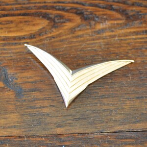 May include: A gold-colored metal brooch in the shape of a stylized bird with a pointed beak and a series of parallel lines running along the body.