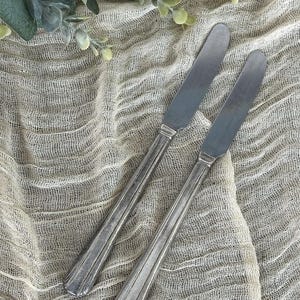 May include: Two silver butter knives with a simple, elegant design. The knives are placed on a textured, off-white fabric, with green foliage in the background. The knives have a rectangular blade and a long, ridged handle.