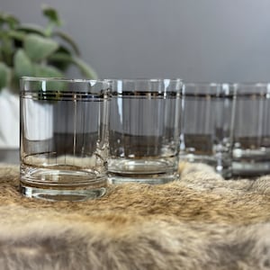 May include: Set of four clear glass tumblers with a gold and silver band around the top. The glasses are sitting on a brown faux fur surface.