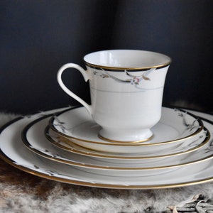 May include: A set of white porcelain dinnerware with a black and gold trim and a floral design. The set includes a cup, saucer, and three plates.