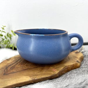 以下が含まれることがあります： ハンドルと注ぎ口が付いた青いセラミックのグレイビーボート。木製のサービングボードに置かれています。ボートの縁には茶色のトリムがあります。背景には緑とフェイクファーのスローが含まれています。