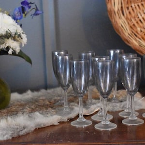 Puede incluir: Un conjunto de copas de champán transparentes con un tinte gris, dispuestas sobre una superficie de madera. Un jarrón con flores blancas y una cesta tejida se encuentran en el fondo. Las copas tienen un diseño clásico.