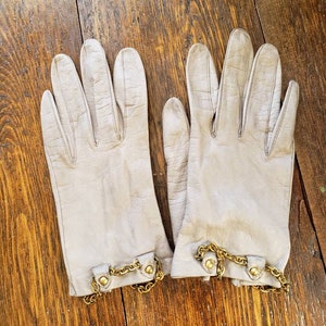 May include: A pair of light gray leather gloves with gold-colored chain and button accents. The gloves are laid out on a wooden surface. The gloves are a classic accessory.