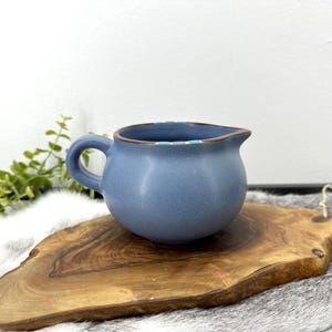 以下が含まれることがあります： 丸みを帯びたボディと小さなハンドルが付いた、青いセラミッククリーマー。 クリーマーには注ぎやすい注ぎ口があり、木製のサービングボードに置かれています。 クリーマーの縁には装飾的な模様があります。