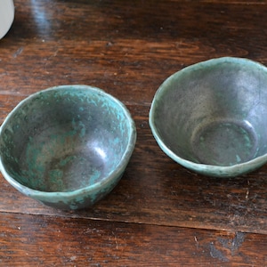 May include: Two handmade ceramic bowls with a textured, turquoise and dark grey finish. The bowls have a rustic, organic shape and are displayed on a dark wooden surface. These bowls are suitable for serving food or as decorative pieces.
