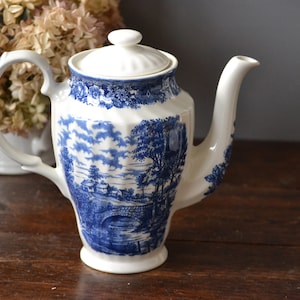 Puede incluir: Una tetera de cerámica blanca con un patrón floral azul y blanco. La tetera tiene una tapa y un asa. La tetera está sentada en una mesa de madera.