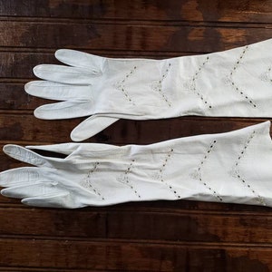 May include: A pair of long, white gloves with decorative cut-out patterns. The gloves are made of a soft material and are laid out on a dark wooden surface. The gloves have a vintage aesthetic.