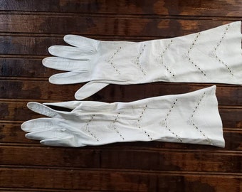Könnte beinhalten: Ein Paar lange, weiße Handschuhe mit dekorativen Ausschnittmustern. Die Handschuhe sind aus einem weichen Material gefertigt und liegen auf einer dunklen Holzoberfläche. Die Handschuhe haben eine Vintage-Ästhetik.