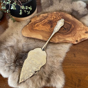 Puede incluir: Utensilio de servir plateado con un diseño floral y las palabras "Merry Christmas" grabadas en la hoja. El utensilio está sobre una superficie de piel sintética marrón.