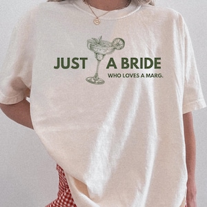 May include: Cream-colored t-shirt with the text "JUST A BRIDE WHO LOVES A MARG." and a margarita glass illustration in green. The shirt has short sleeves and a relaxed fit. The bottom of the shirt is paired with red and white checkered shorts.