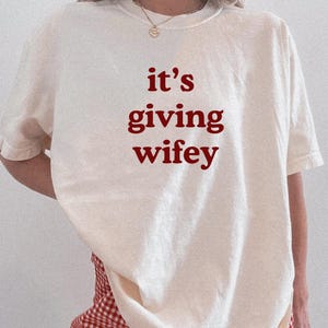 Może przedstawiać: Kremowa koszulka z napisem "it's giving wifey" w bordowym kolorze. Koszulka jest zestawiona z czerwono-białymi szortami w kratę. Tekst "White shirt" i "Chili print" znajduje się na dole obrazu.