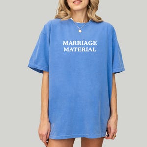 Op de afbeelding: Een blauw t-shirt met de woorden "MARRIAGE MATERIAL" in het wit gedrukt. Het shirt heeft korte mouwen en een relaxte pasvorm. Het model draagt een gouden ketting.