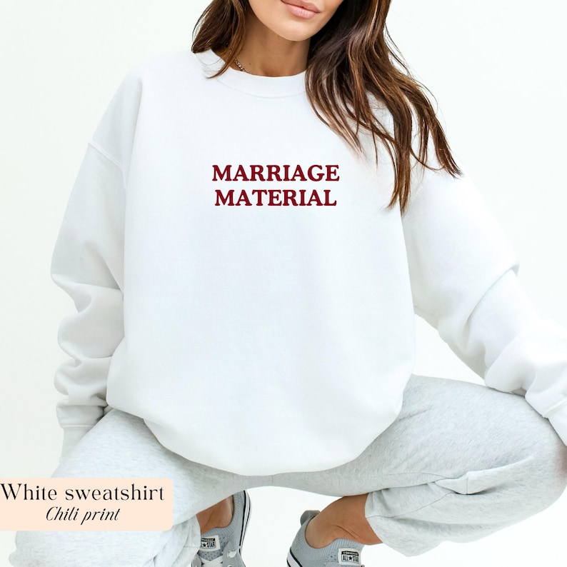 Op de afbeelding: Een witte sweatshirt met de tekst "MARRIAGE MATERIAL" in bordeauxrood. De sweatshirt heeft een ronde hals en lijkt gemaakt te zijn van een zacht materiaal. De afbeelding bevat ook de tekst "White sweatshirt Chili print".