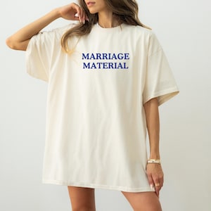 Op de afbeelding: Cr&egrave;mekleurig, oversized T-shirt met de woorden "MARRIAGE MATERIAL" in blauw gedrukt. Het shirt heeft korte mouwen en een relaxte pasvorm. De tekst staat in het midden van de voorkant van het shirt.
