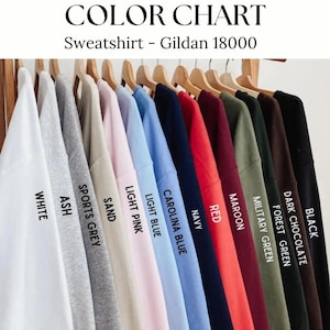 Op de afbeelding: Een kleurenkaart met een verscheidenheid aan sweatshirtkleuren, waaronder wit, as, sportgrijs, zand, lichtroze, lichtblauw, Carolina blauw, marine, rood, bordeaux, legergroen, bosgroen, donkere chocolade en zwart. De tekst luidt "Sweatshirt - Gildan 18000".