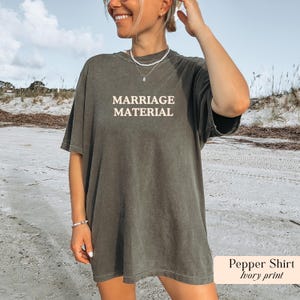 Op de afbeelding: Een donker olijfgroen t-shirt met de tekst "MARRIAGE MATERIAL" in licht ivoor. Het shirt heeft een relaxte pasvorm en lijkt gemaakt te zijn van een zacht materiaal. Het shirt maakt deel uit van de "Pepper Shirt" collectie.