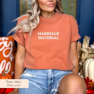 Op de afbeelding: Een verbrand oranje t-shirt met de woorden "MARRIAGE MATERIAL" in het wit gedrukt. Het shirt heeft korte mouwen en een relaxte pasvorm. Het model draagt een spijkerbroek.