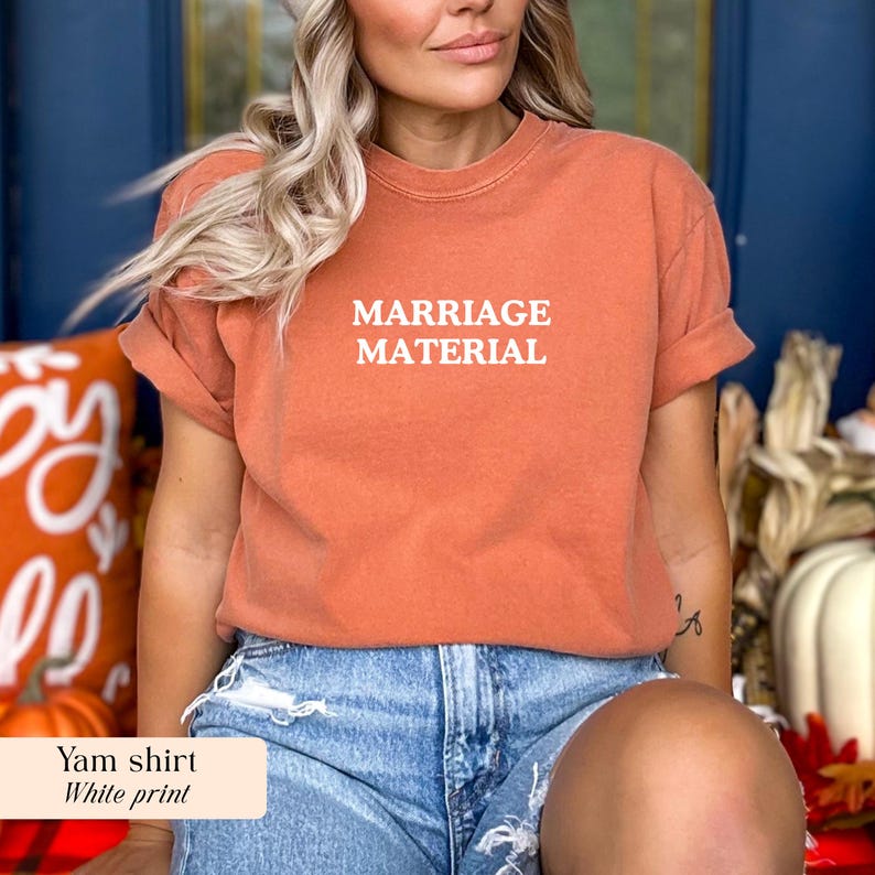 Op de afbeelding: Een verbrand oranje t-shirt met de woorden "MARRIAGE MATERIAL" in het wit gedrukt. Het shirt heeft korte mouwen en een relaxte pasvorm. Het model draagt een spijkerbroek.
