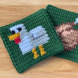 Puede incluir: Dos salvamanteles cuadrados de ganchillo verde. Uno presenta un pollo de dibujos animados con detalles blancos, grises, amarillos y rojos. El otro tiene un diseño de pavo marrón con detalles azules. Sobre una superficie de madera.