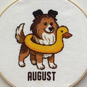 Puede incluir: Bordado de punto de cruz de un perro con un flotador amarillo con forma de pato. El perro es marrón, blanco y negro, con la palabra "AGOSTO" bordada debajo. El bordado está en un aro de madera.