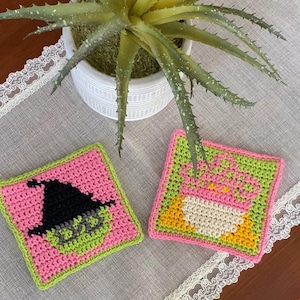 Könnte beinhalten: Zwei quadratische Häkeluntersetzer. Einer zeigt ein Hexengesicht und einen Hut in Schwarz und Grün auf rosa Hintergrund. Der andere Untersetzer hat ein Cupcake-Design in Weiß, Gelb und Grün auf rosa Hintergrund. Beide haben grüne und rosa Ränder.