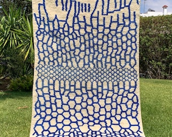 Puede incluir: Una alfombra rectangular de color crema con un patrón geométrico azul. El diseño presenta una serie de formas y líneas interconectadas, creando una apariencia texturizada. La alfombra se exhibe al aire libre sobre hierba verde, con flecos en el borde inferior.