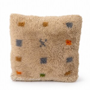 Peut inclure: Un coussin carré et moelleux de couleur beige, orné d'un motif de petits carrés colorés et d'une figure stylisée. Les carrés sont dans des tons orange, vert, gris et marron. Le texte "The Rug Caravane" est en bas.