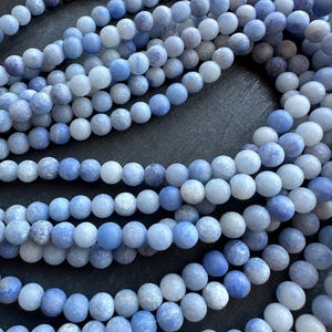 May include: Close-up of several strands of blue gemstone beads. The beads are a mix of light and medium blue hues, with a matte finish. The round beads are strung together, creating a textured and visually appealing pattern. Ideal for jewelry making.