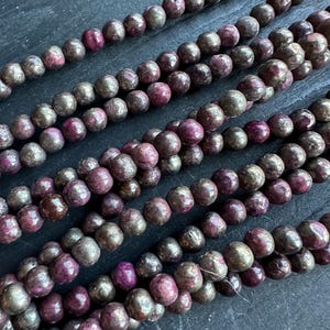 May include: Strands of round beads in shades of burgundy, purple, and bronze. The beads have a metallic sheen and are strung together, creating a textured and colorful display. Ideal for jewelry making.