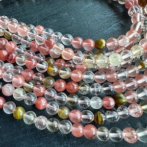 May include: Strands of gemstone beads in varying shades of pink, red, yellow, and clear. The beads are round and appear to be strung together, creating a textured and colorful display. The beads are likely used for jewelry making.