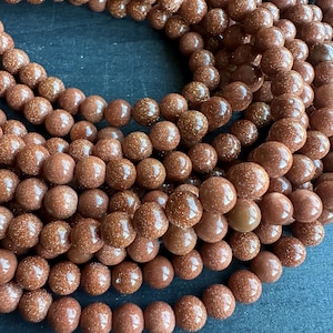 May include: Close-up of strands of brown, round beads with a sparkling, glittery appearance. The beads are a warm, earthy tone, and the image showcases the texture and detail of the gemstone beads. Ideal for jewelry making.