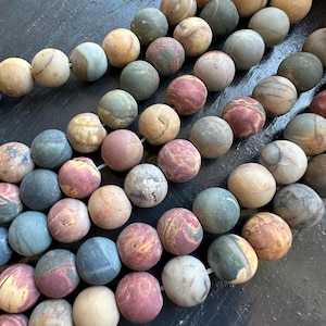 May include: Strands of round gemstone beads in various colors, including beige, pink, blue, and green. The beads have a matte finish and natural variations in color and pattern, suggesting they are made of stone.