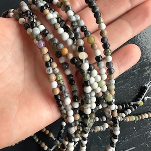 May include: Strands of small, round beads in various colors including white, black, gray, brown, and green. The beads are strung together, creating a textured and colorful display. The beads are likely for jewelry making.