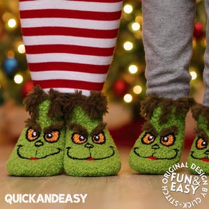 Könnte beinhalten: Grüne Grinch-Slipper mit gestickten Augen, Augenbrauen und einem lächelnden Mund. Die Slipper haben braune Fellakzente und werden mit rot-weiß gestreiften Socken und grauen Hosen getragen. Der Text "QUICK AND EASY" ist sichtbar.
