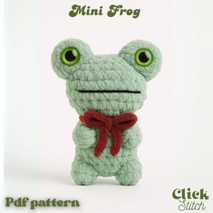 May include: A light green crocheted mini frog with large, round eyes and a red bow. The frog has a black stitched mouth and is standing upright. Text on the image reads "Mini Frog" and "Pdf pattern".