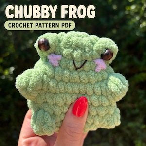 May include: A light green crochet frog plushie with dark brown bead eyes and embroidered pink cheeks. The text CHUBBY FROG CROCHET PATTERN PDF is displayed at the top. The frog has a rounded body, small arms, and a stitched mouth.