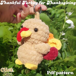 May include: A plush turkey keychain in shades of tan, red, yellow, and orange. The turkey has a fluffy body, a red wattle, and a yellow beak. The keychain is held by a hand against a backdrop of green foliage. Text reads "Thankful Turkey for Thanksgiving".