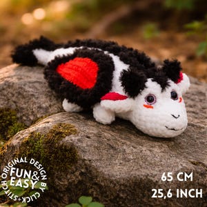 May include: A handmade crochet creature toy in black, white, and red, resting on a rock. The toy has a heart-shaped red marking and measures 25.6 inches long. The text "FUN & EASY" is visible.