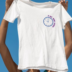 May include: White t-shirt with a round clock graphic and the text "If not now, when?" in purple. The clock has a blue border and black hands. The shirt is held up against a bright blue sky.