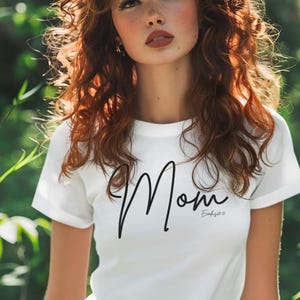 Puede incluir: Camiseta blanca con la palabra "Mom" en una escritura elegante. La camiseta se usa con pantalones verdes. La imagen está tomada al aire libre con un fondo verde borroso.