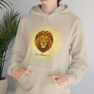 Puede incluir: Sudadera con capucha beige con un diseño de cabeza de león y un halo dorado. El texto "Revelation 5:5" está impreso debajo. La sudadera tiene un bolsillo de canguro y una capucha con cordón.