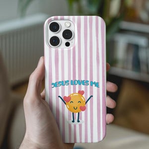 Puede incluir: Funda de teléfono con un diseño de rayas rosas y blancas. La funda presenta el texto "JESUS LOVES ME" y un personaje amarillo sonriente con corazones. La funda del teléfono está en la mano de una persona.