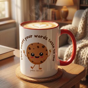 Puede incluir: Una taza de cerámica blanca con borde y asa rojos, llena de café con arte latte en forma de corazón. La taza muestra un personaje de galleta con chispas de chocolate y el texto: How sweet your words taste to me, psalm 119:103.