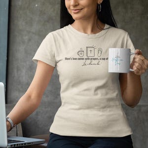 Puede incluir: Una camiseta color crema con el texto "Mom's love comes with prayers, a cup of La Chancla" y dibujos de una taza de café, un libro y sandalias. Una taza blanca con texto azul se sostiene en la mano derecha.