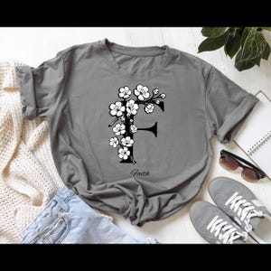 Puede incluir: Una camiseta gris con un diseño floral "F" blanco y la palabra "Faith" impresa debajo. La camiseta se combina con vaqueros azules, zapatillas grises y gafas de sol.