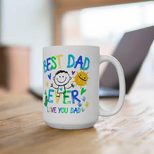 Puede incluir: Una taza de cerámica blanca con un diseño gráfico colorido que incluye el texto BEST DAD EVER y LOVE YOU DAD. La ilustración presenta una figura de palitos, un sol amarillo, corazones pequeños y huellas de manos en tinta azul, verde, amarilla y morada.