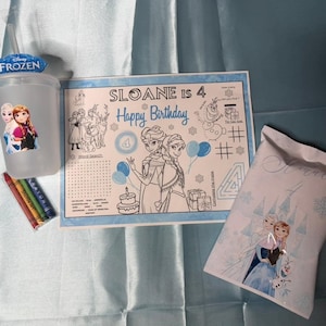 May include: A clear plastic cup with a Frozen logo lid and straw, three crayons, a rectangular activity sheet with the text Sloane is 4 Happy Birthday, and a white paper bag featuring an illustration of Elsa, Anna, and Olaf from Frozen.
